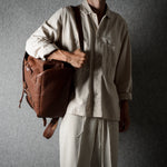A person wearing a light beige button-up shirt and matching pants carries a brown leather duffel bag over their shoulder against a dark gray textured background.