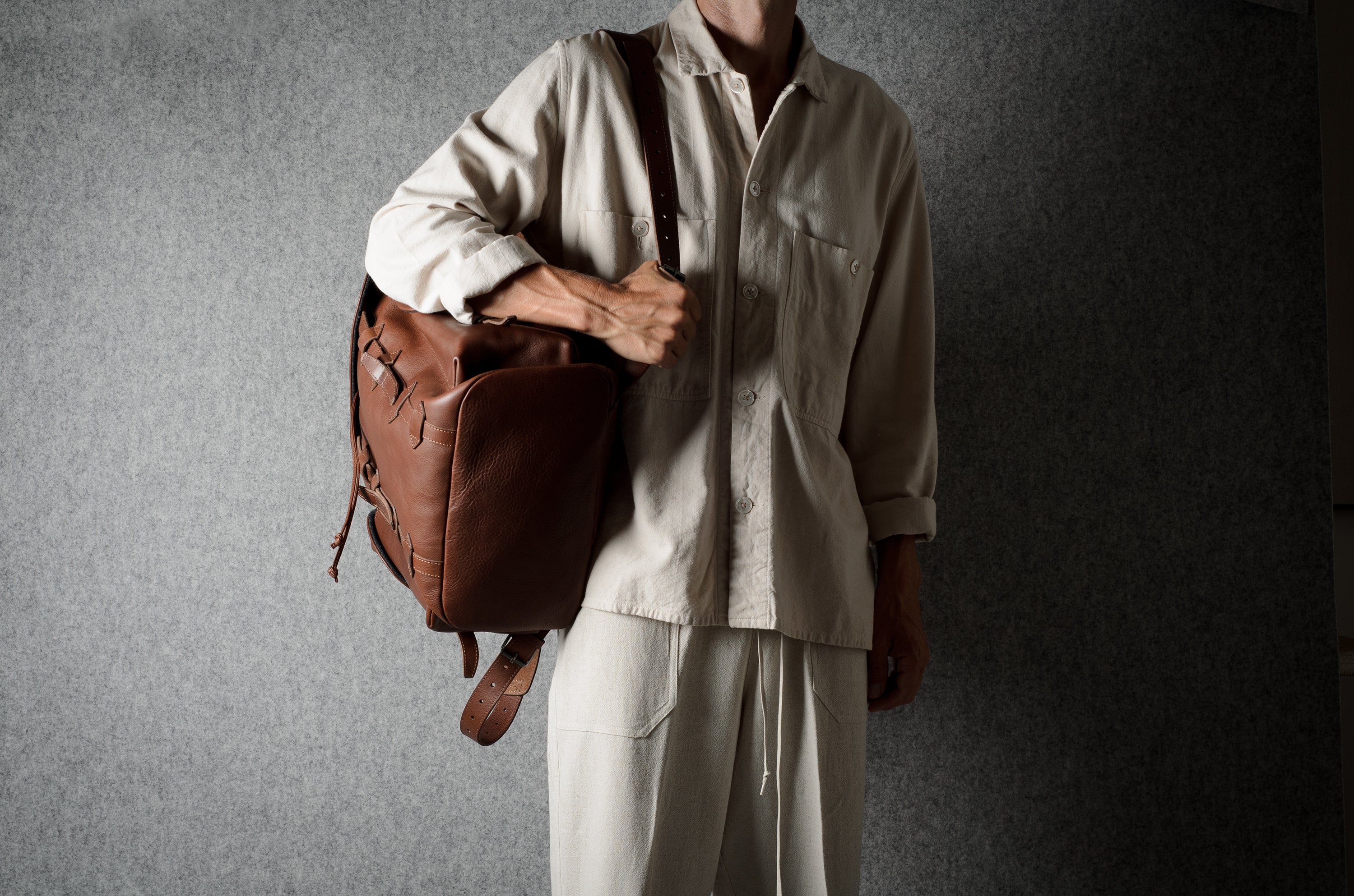 A person wearing a light beige button-up shirt and matching pants carries a brown leather duffel bag over their shoulder against a dark gray textured background.