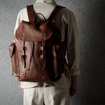 A person viewed from behind wearing a brown leather backpack with multiple front straps and side pockets, standing against a dark textured gray wall.