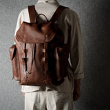 A person viewed from behind wearing a brown leather backpack with multiple front straps and side pockets, standing against a dark textured gray wall.