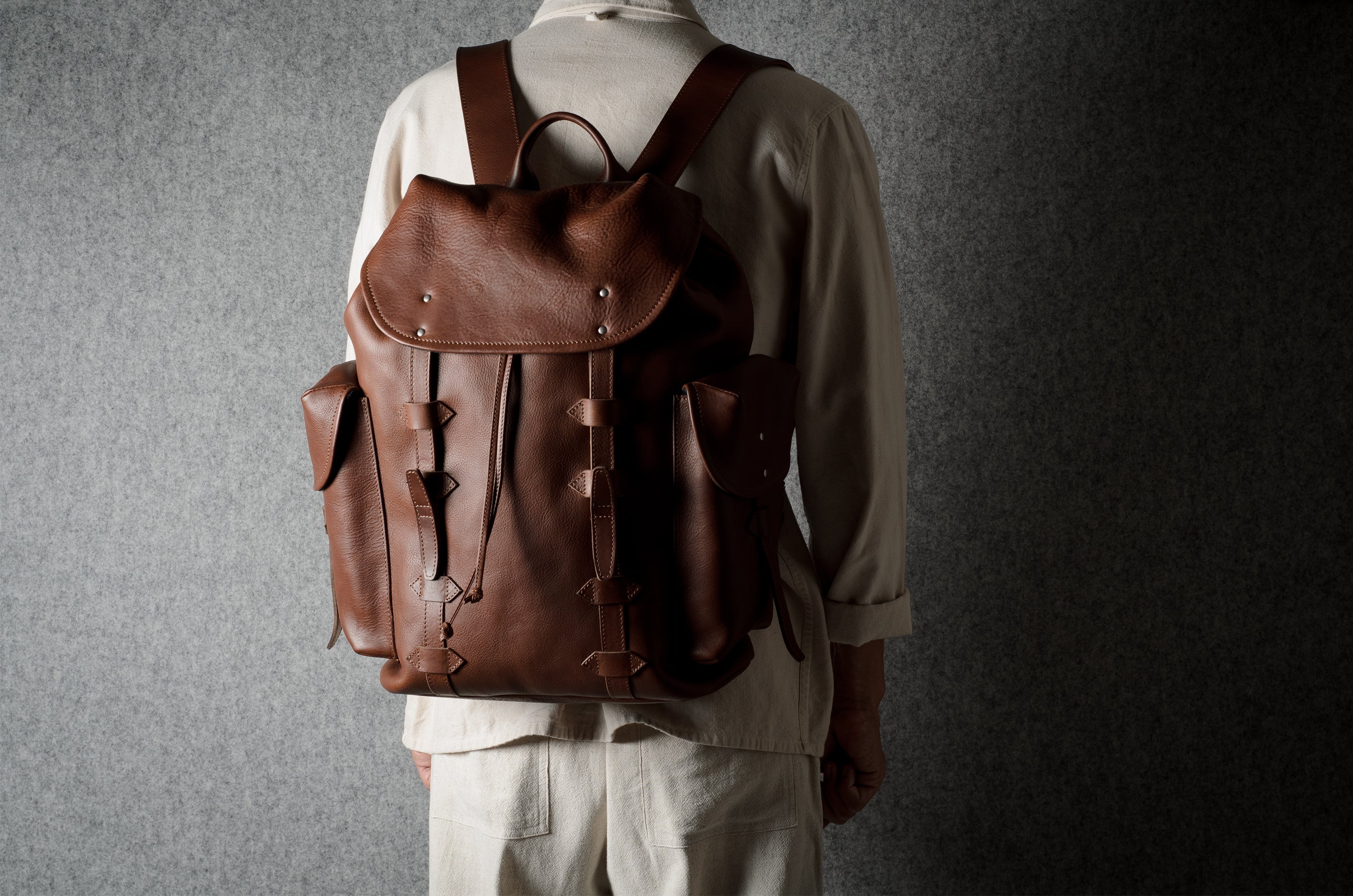 A person viewed from behind wearing a brown leather backpack with multiple front straps and side pockets, standing against a dark textured gray wall.
