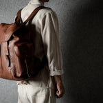 A person wearing a light beige shirt and trousers, shown from the back and side, carrying a brown leather backpack against a dark textured gray wall.