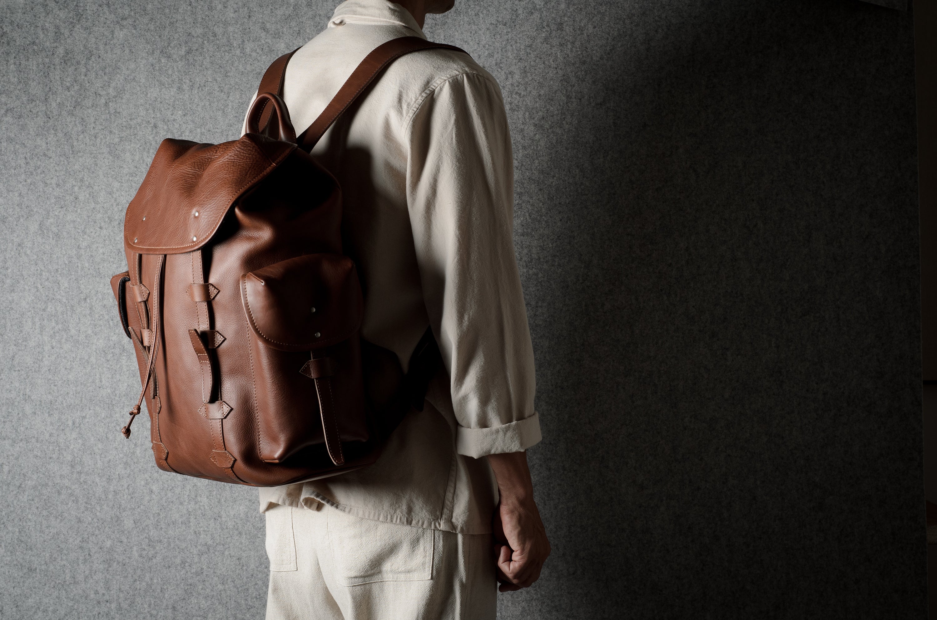 A person wearing a light beige shirt and trousers, shown from the back and side, carrying a brown leather backpack against a dark textured gray wall.