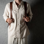 A person wearing a cream-colored linen shirt and matching trousers, holding a brown leather crossbody bag with brown leather shoulder straps against a dark gray textured background.