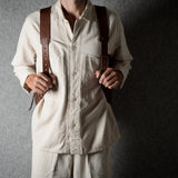 A person wearing a cream-colored linen shirt and matching trousers, holding a brown leather crossbody bag with brown leather shoulder straps against a dark gray textured background.
