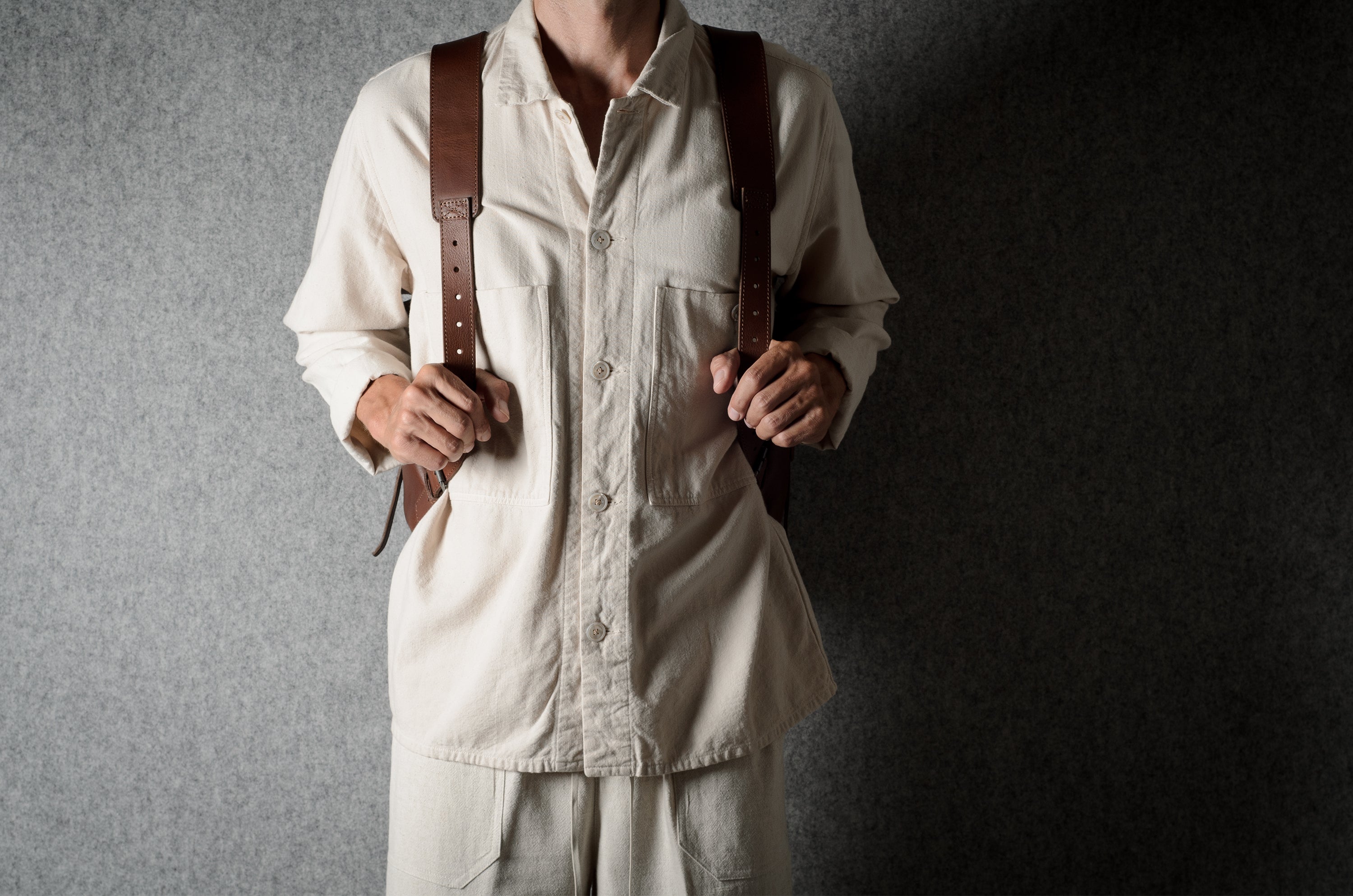 A person wearing a cream-colored linen shirt and matching trousers, holding a brown leather crossbody bag with brown leather shoulder straps against a dark gray textured background.