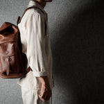 A person in a light beige shirt and pants wearing a brown leather backpack, shown from the side against a textured gray wall with dramatic lighting.