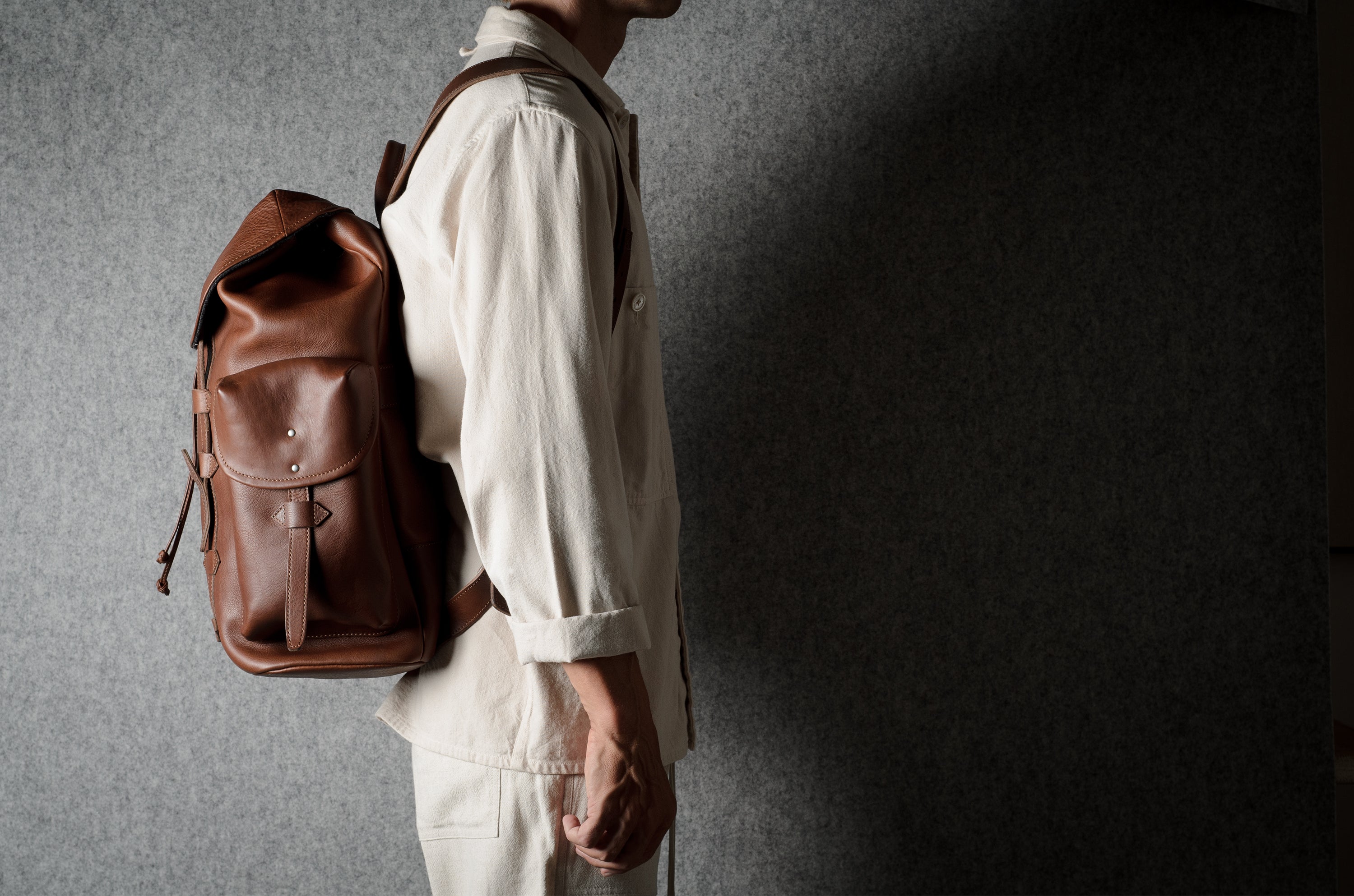 A person in a light beige shirt and pants wearing a brown leather backpack, shown from the side against a textured gray wall with dramatic lighting.