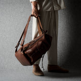 A person in light beige clothing holds a brown leather duffel bag with a long shoulder strap against a dark gray textured studio backdrop.
