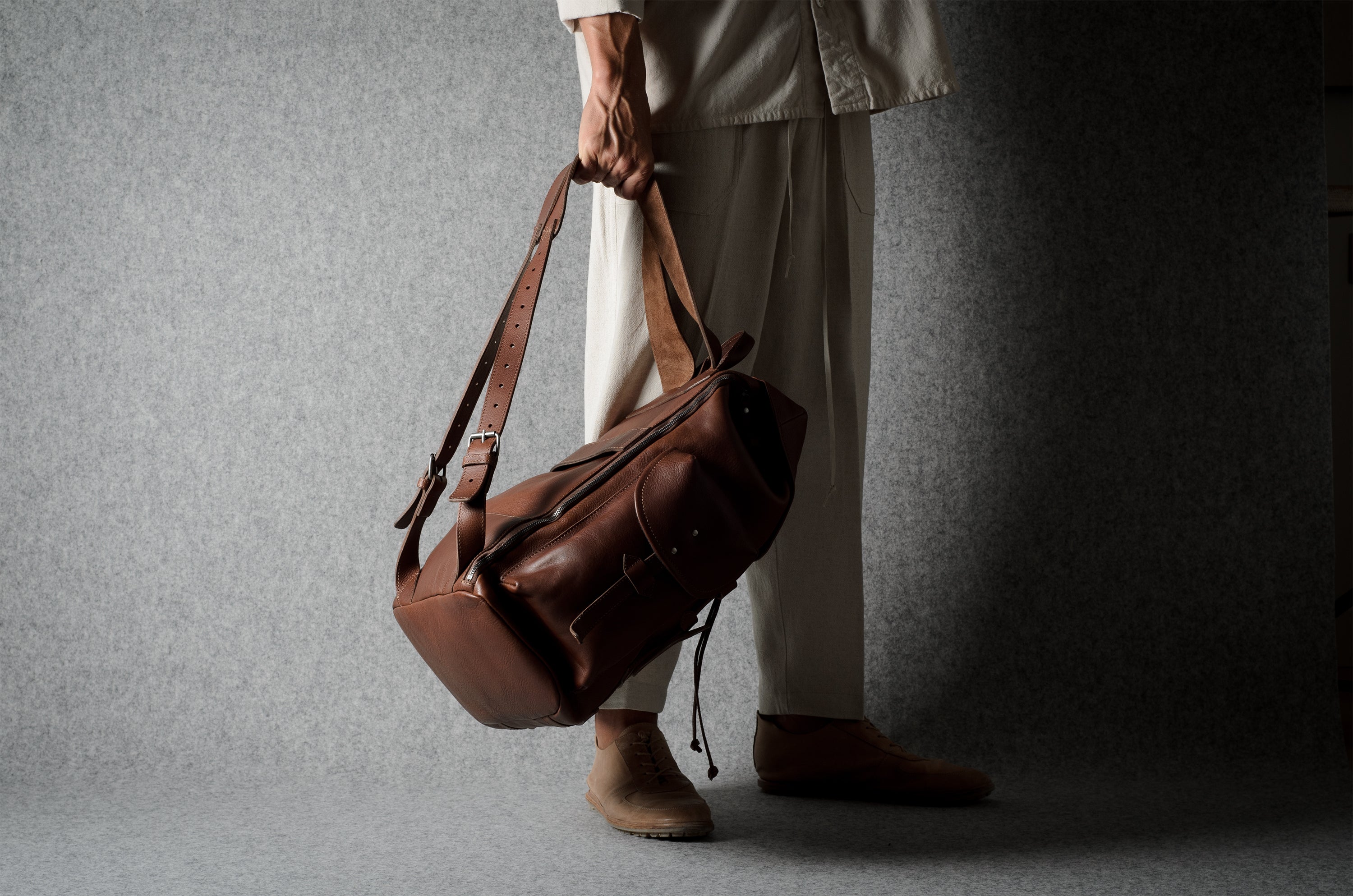 A person in light beige clothing holds a brown leather duffel bag with a long shoulder strap against a dark gray textured studio backdrop.