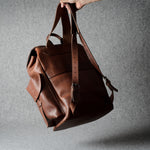 A brown leather backpack hanging from a hand against a textured gray studio background.