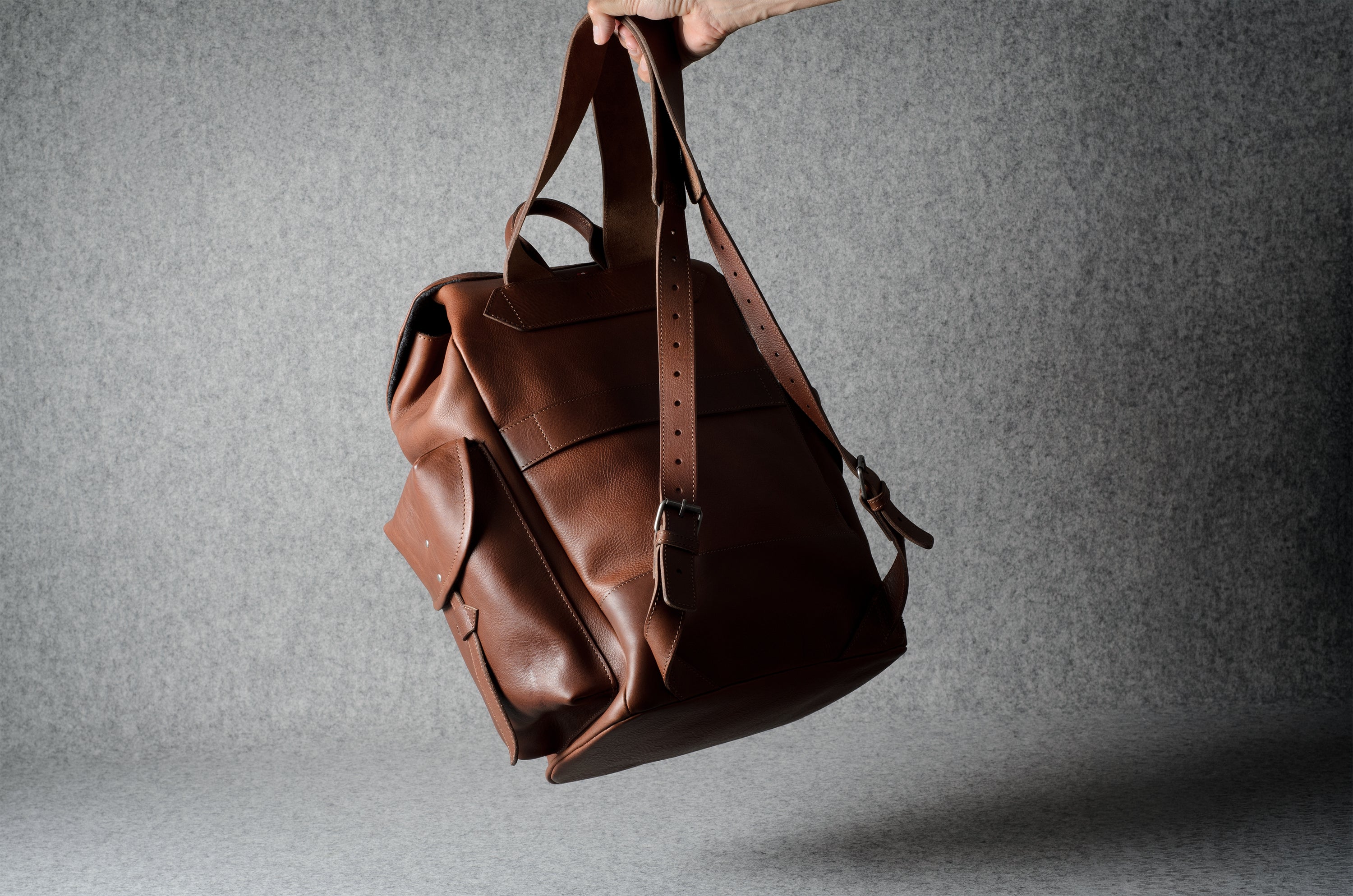 A brown leather backpack hanging from a hand against a textured gray studio background.