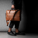 Brown leather briefcase being carried by a man wearing grey on a grey background.