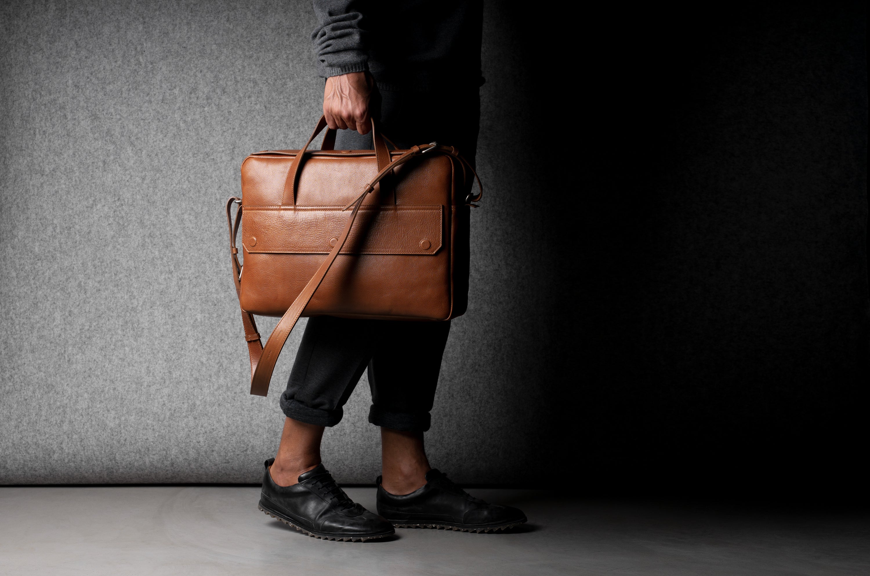 Brown leather briefcase being carried by a man wearing grey on a grey background.