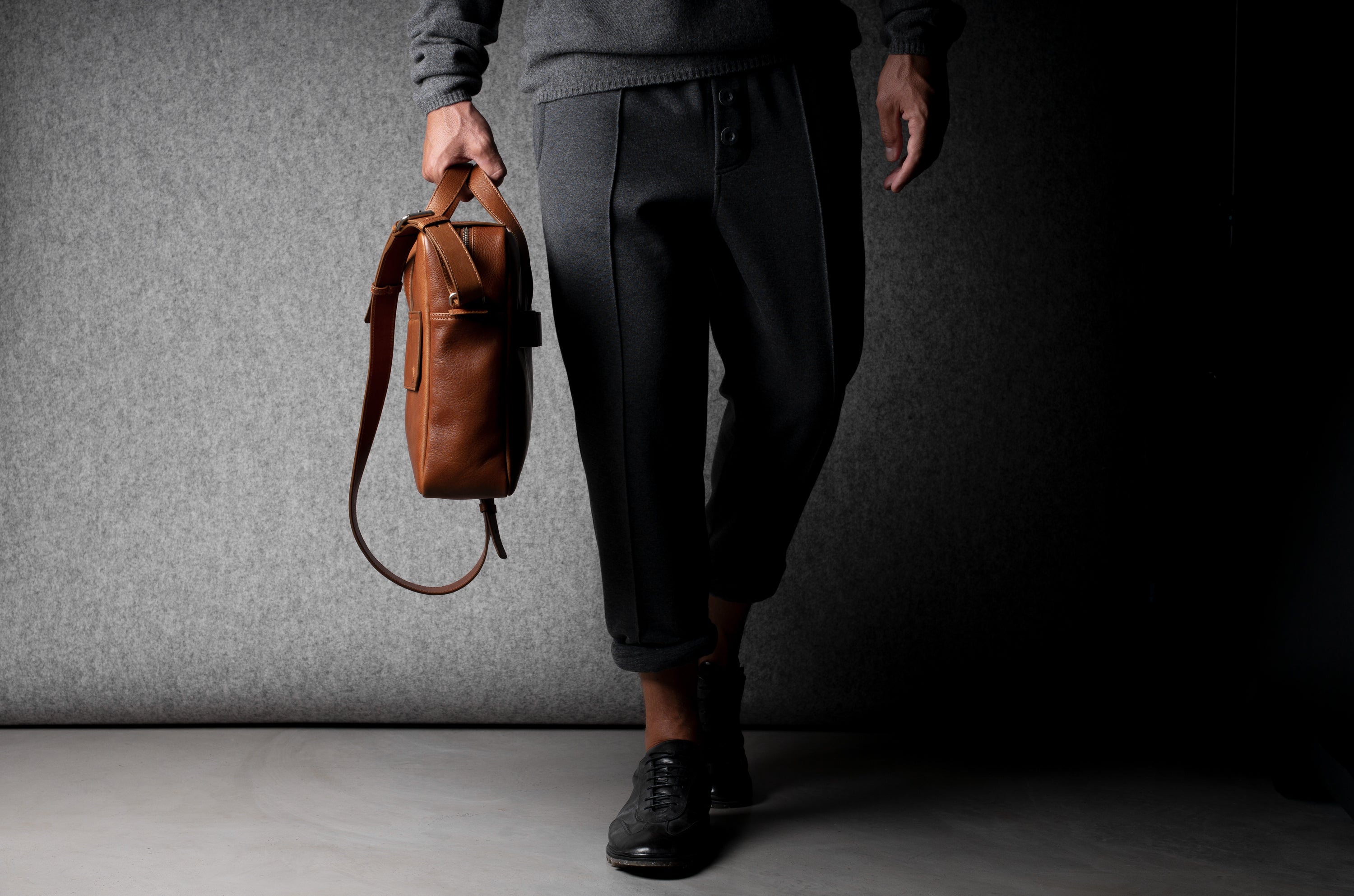 Brown leather briefcase being carried by a man wearing grey on a grey background.
