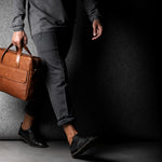 Brown leather briefcase being carried by a man wearing grey on a grey background.