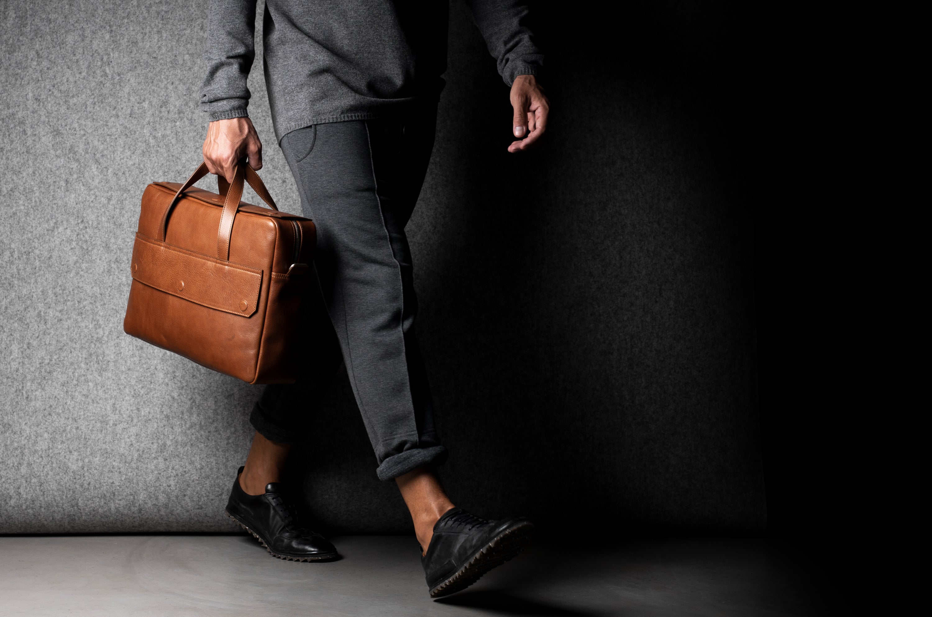 Brown leather briefcase being carried by a man wearing grey on a grey background.