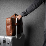 Person holding a brown leather bag over a silver suitcase against a gray background