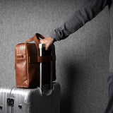 Person holding a brown leather bag over a silver suitcase against a gray background
