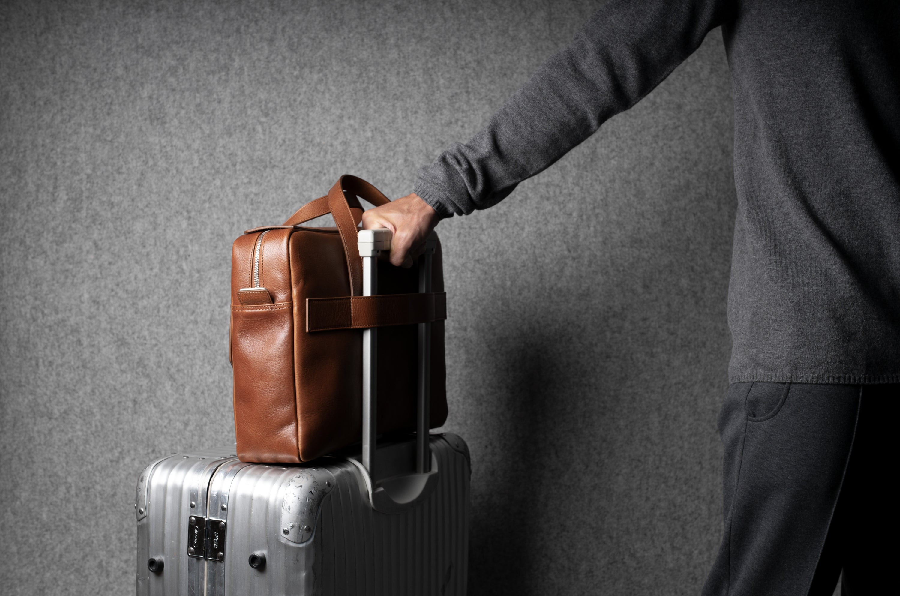Person holding a brown leather bag over a silver suitcase against a gray background