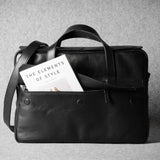 Black leather bag with a book titled 'The Elements of Style' on a gray background