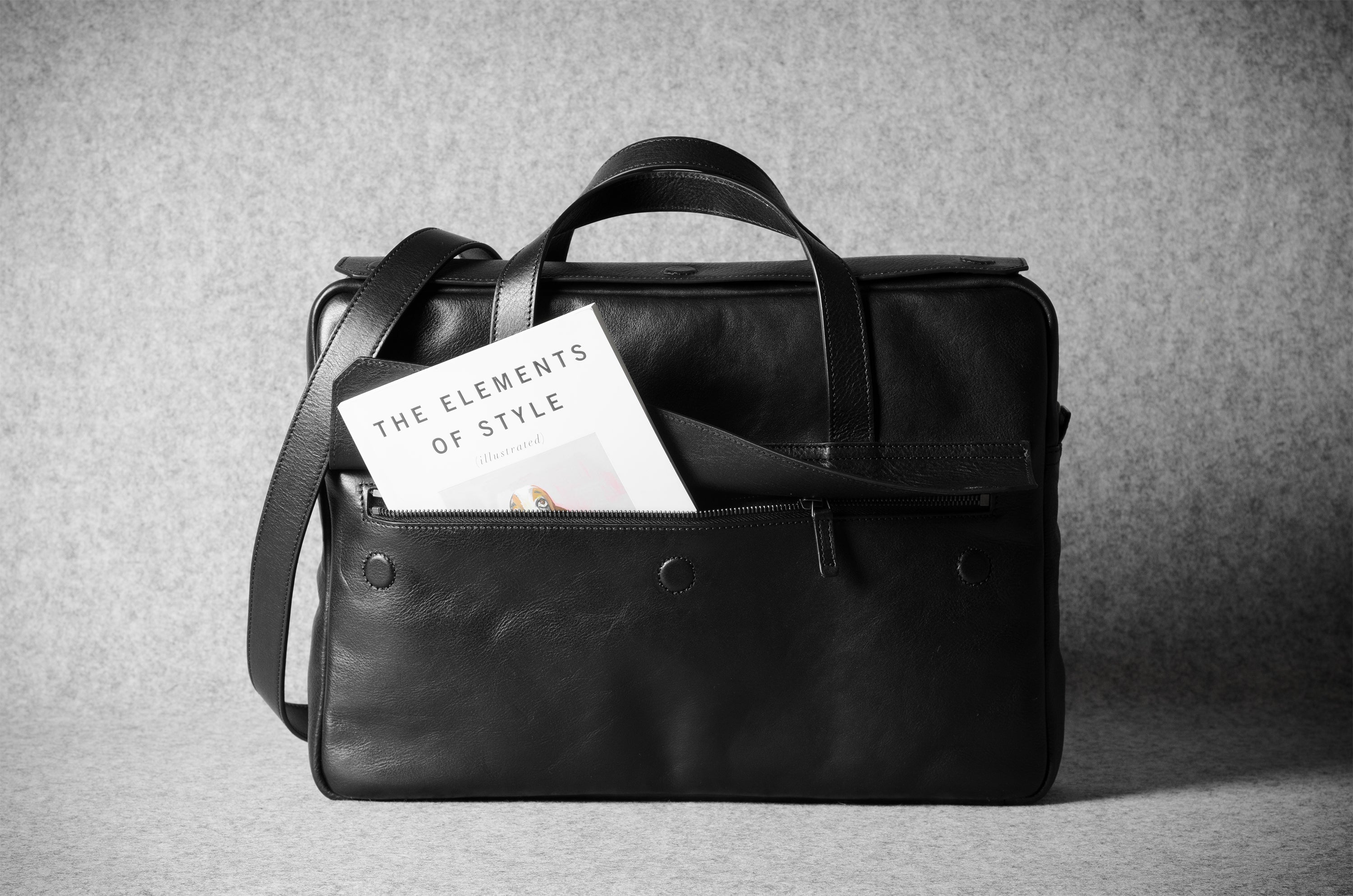 Black leather bag with a book titled 'The Elements of Style' on a gray background