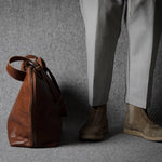 Brown leather bag next to a person wearing light gray pants and brown shoes on a gray background