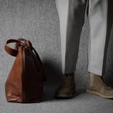 Brown leather bag next to a person wearing light gray pants and brown shoes on a gray background
