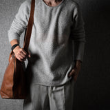 Person wearing a gray wool sweater holding a brown leather bag against a dark background