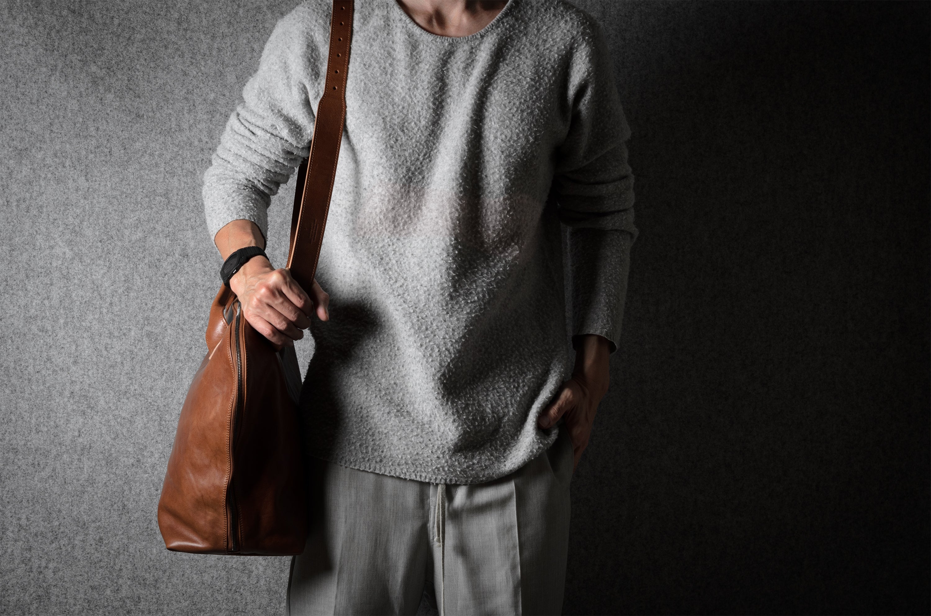 Person wearing a gray wool sweater holding a brown leather bag against a dark background