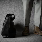 Person wearing light-colored pants and shoes standing next to a black leather bag on a textured gray floor.