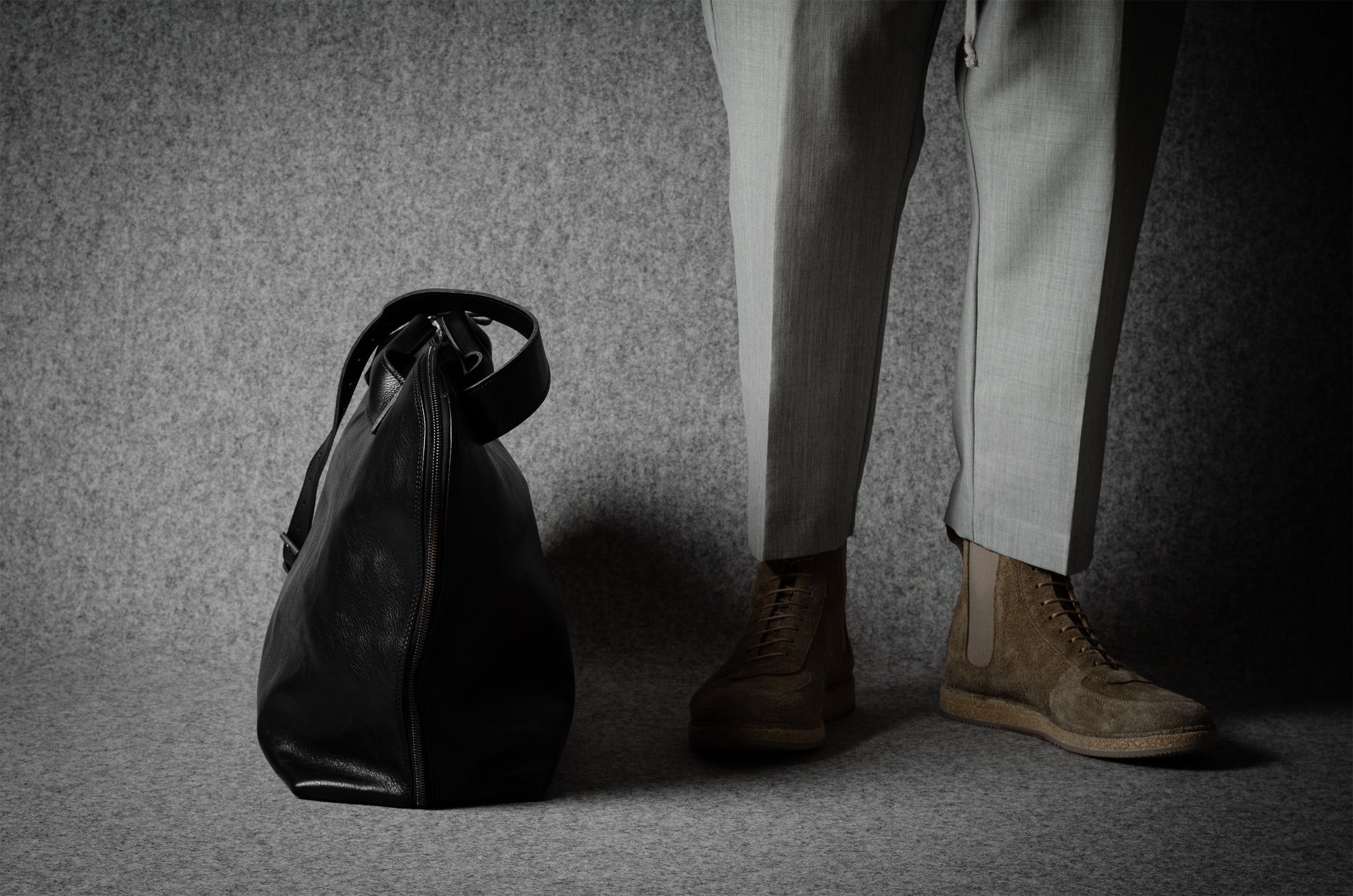 Person wearing light-colored pants and shoes standing next to a black leather bag on a textured gray floor.