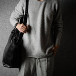 Person wearing a gray sweater and holding a black leather bag against a dark background