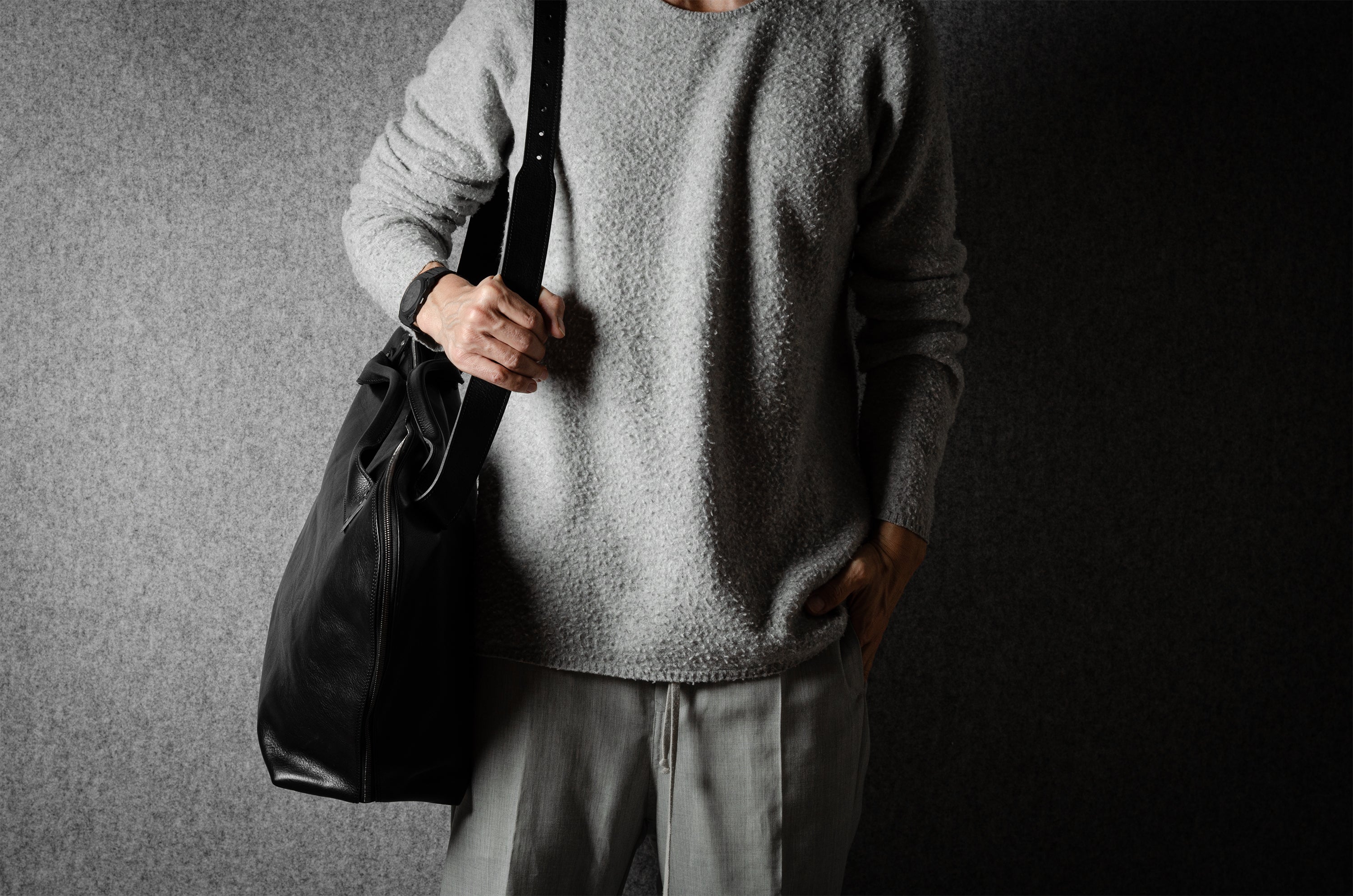 Person wearing a gray sweater and holding a black leather bag against a dark background