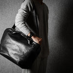 Person holding a black leather duffel bag against a gray background