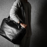 Person holding a black leather duffel bag against a gray background
