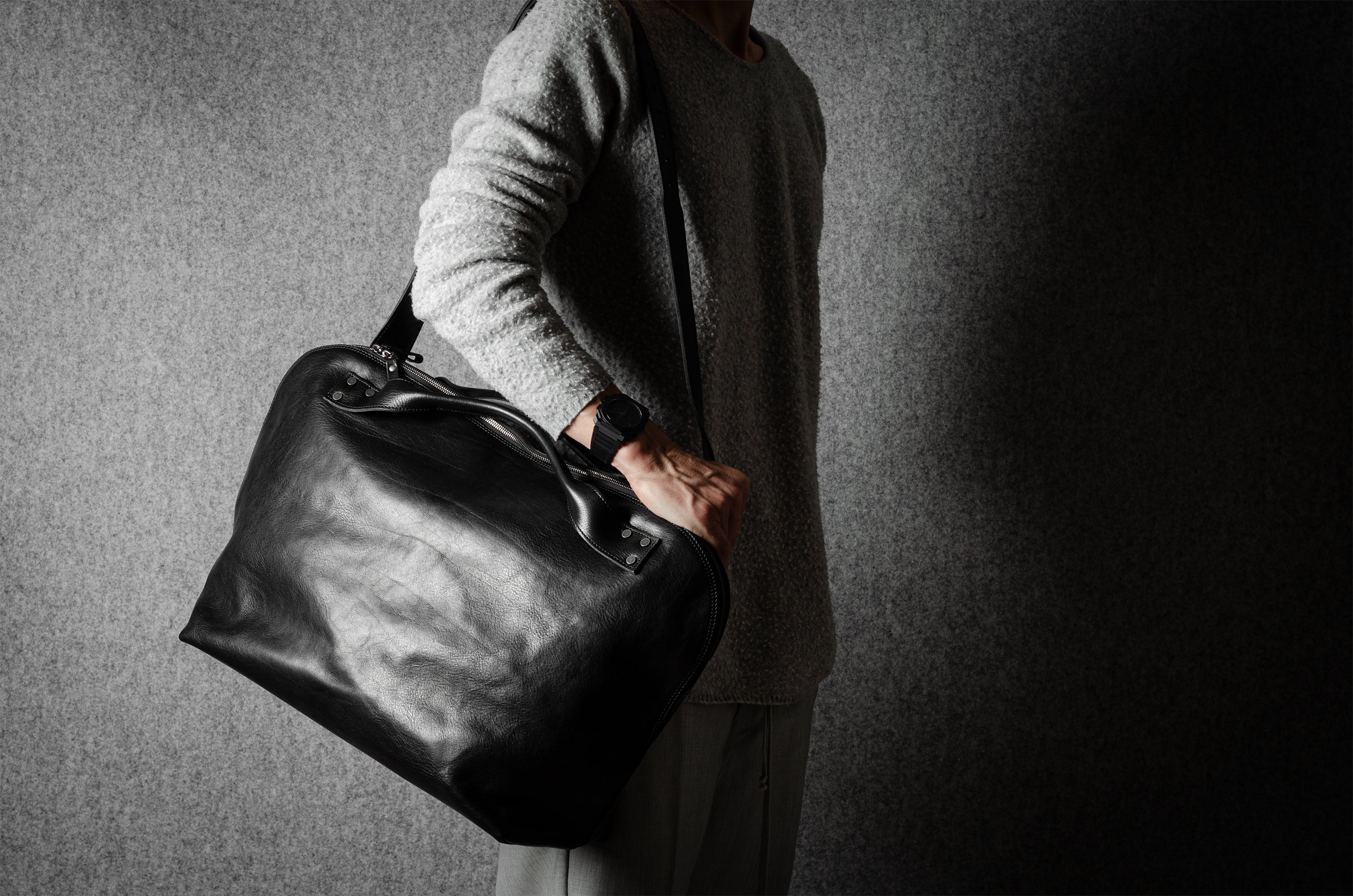 Person holding a black leather duffel bag against a gray background