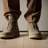 Person wearing brown suede boots and pants on a wooden floor with a neutral background