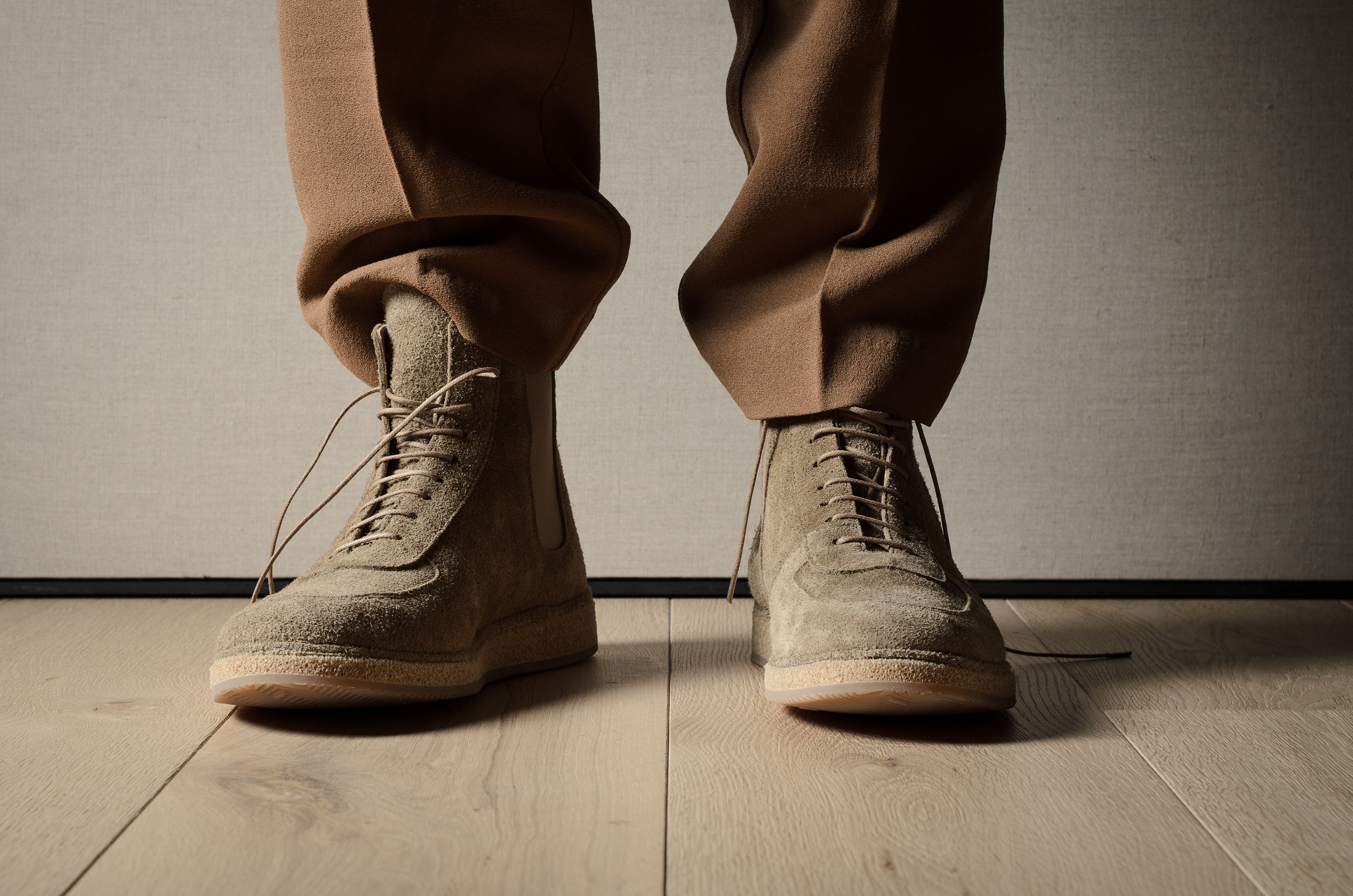 Person wearing brown suede boots and pants on a wooden floor with a neutral background