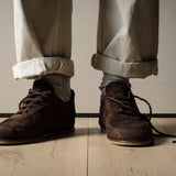 Brown suede shoes worn with rolled-up beige pants on a wooden floor.