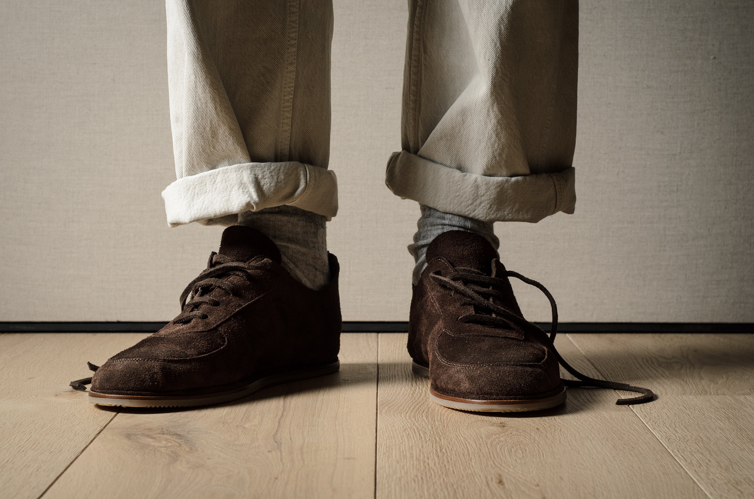 Brown suede shoes worn with rolled-up beige pants on a wooden floor.