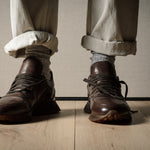 Brown leather sneakers worn with rolled-up beige pants on a wooden floor.