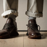 Brown leather sneakers worn with rolled-up beige pants on a wooden floor.