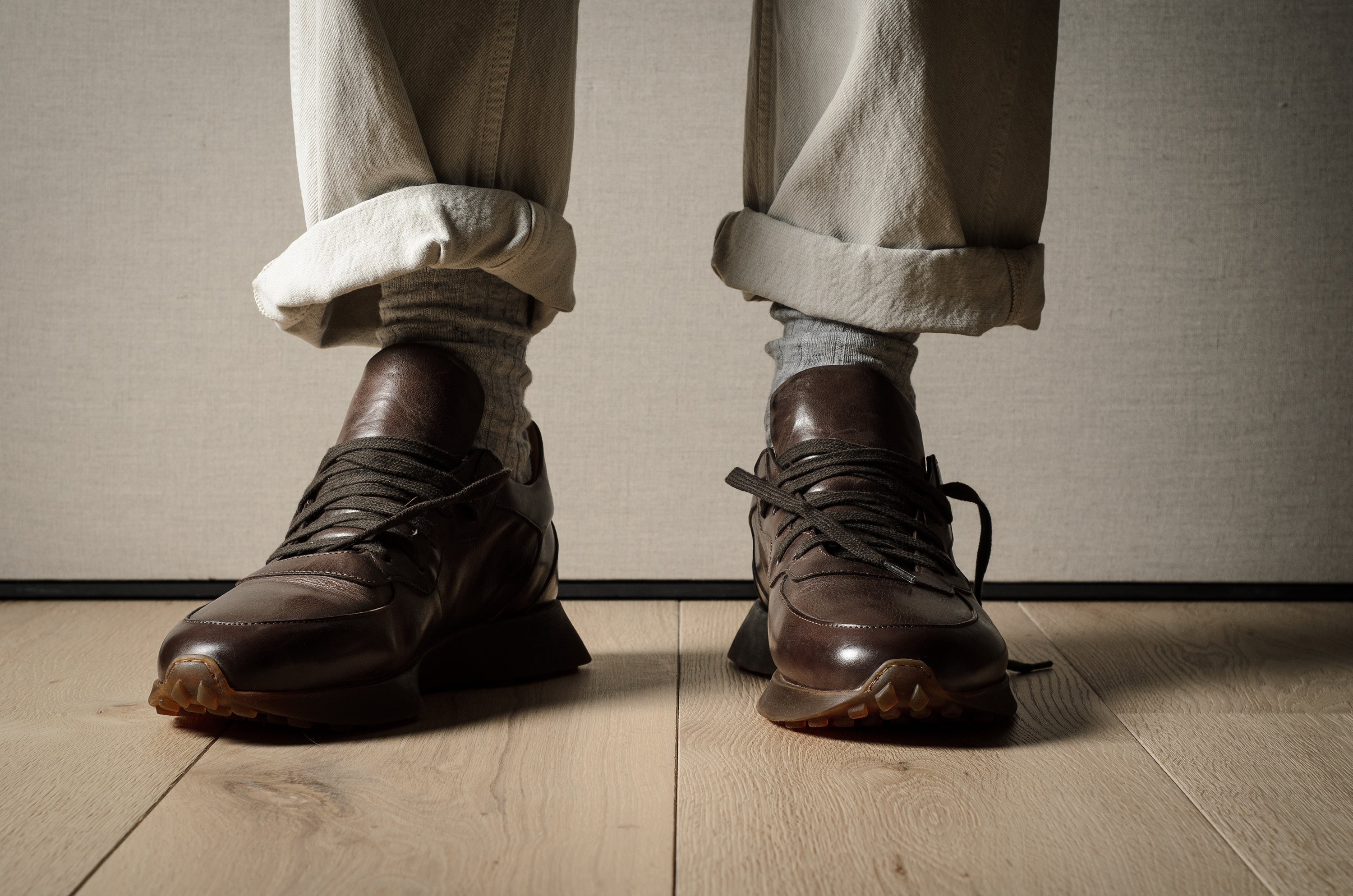 Brown leather sneakers worn with rolled-up beige pants on a wooden floor.