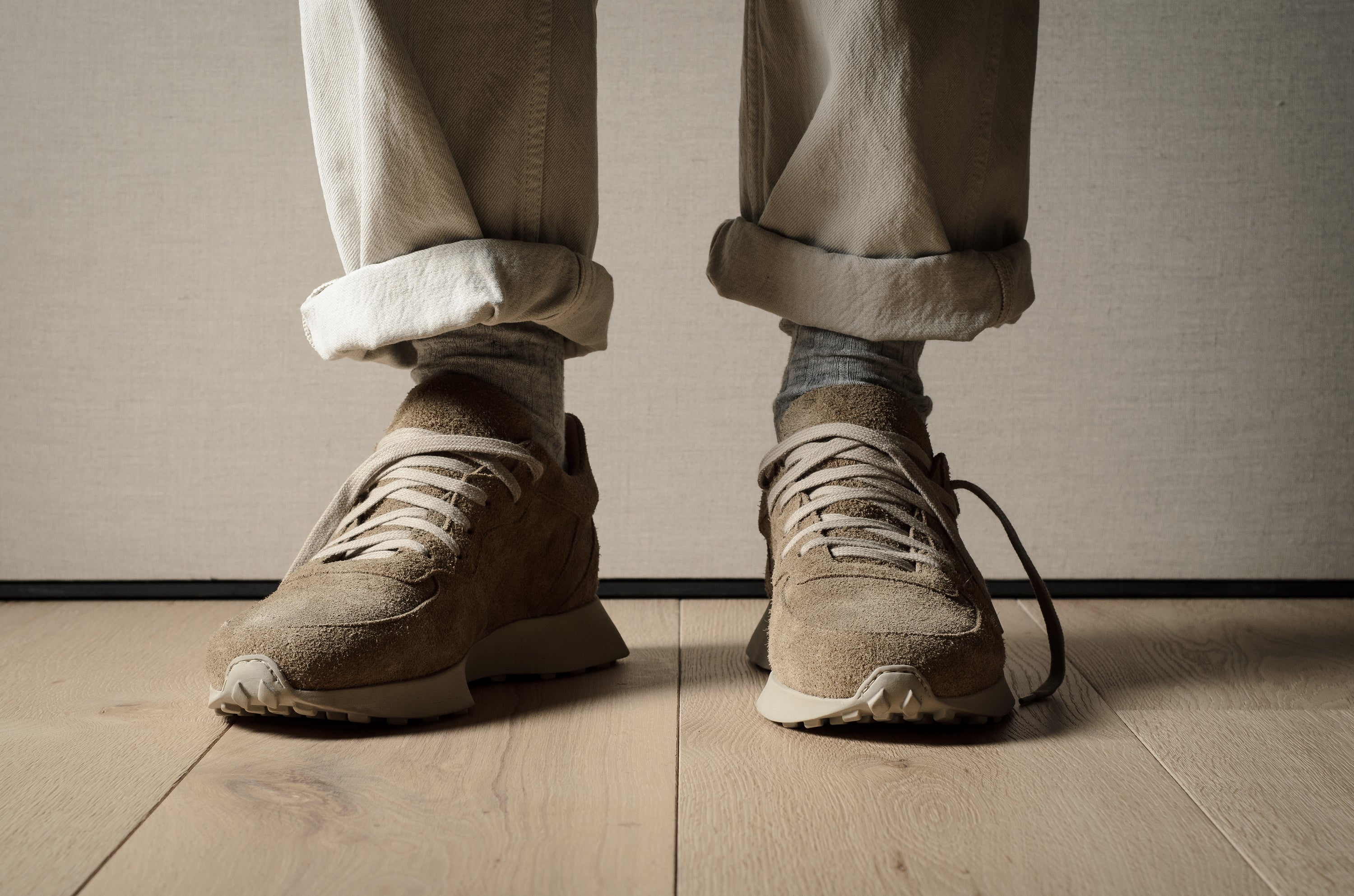 Close-up of a person wearing beige sneakers and rolled-up pants on a wooden floor.