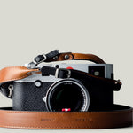 A camera with a brown and black duotone leather strap, designed to hang around the neck.